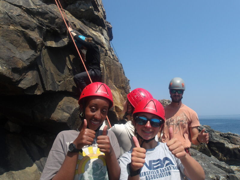 The image shows a group of people rock climbing. Two people in the foreground are smiling and giving a thumbs up. They are wearing helmets and sunglasses. In the background, there is a person climbing a rock face and another person is standing and looking at the camera. The sky is blue and the sun is shining.